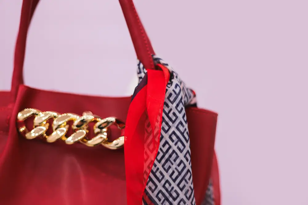 Close-up of a stylish red leather handbag with a gold chain detail, elegantly accessorized with a red and navy patterned scarf, perfect for adding a chic touch to any outfit.